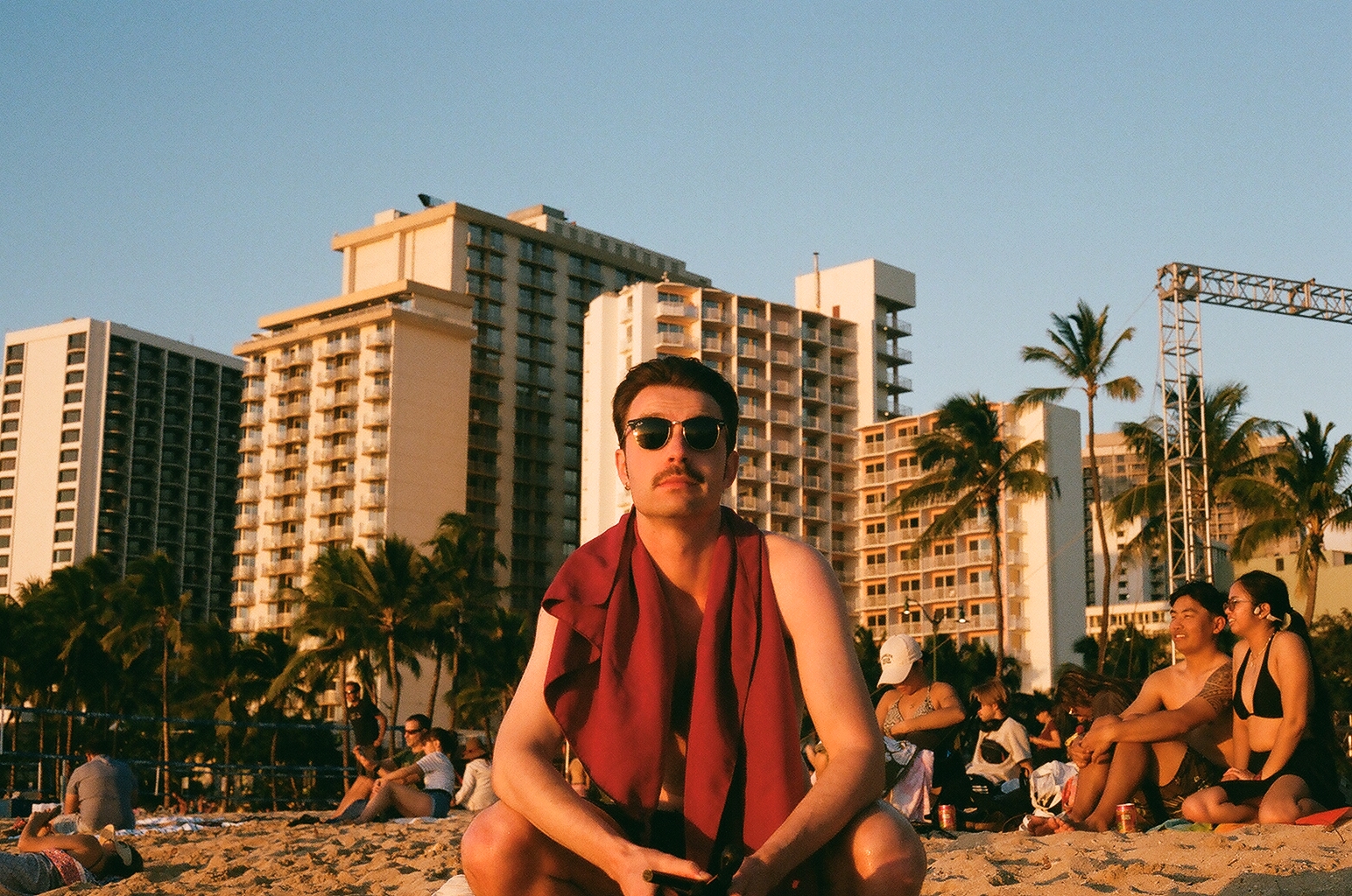 A man in sunglasses with a red towel around his neck, Waikiki hotels behind him in golden light