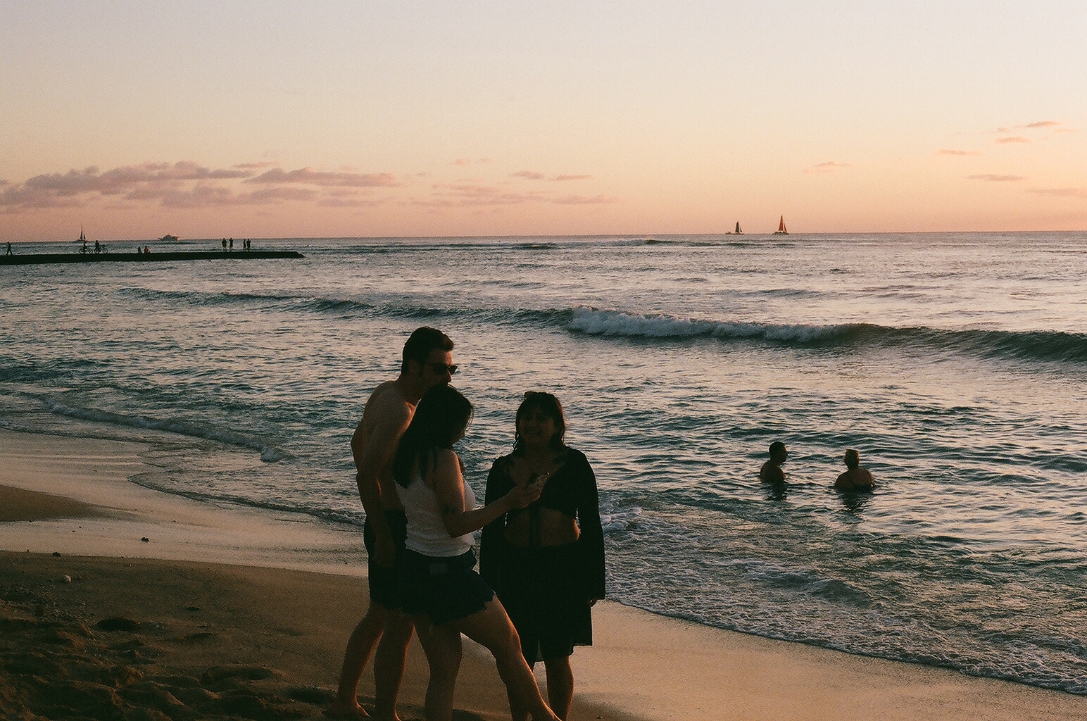 Three friends chat at the shoreline at dusk while swimmers and distant sailboats dot the water