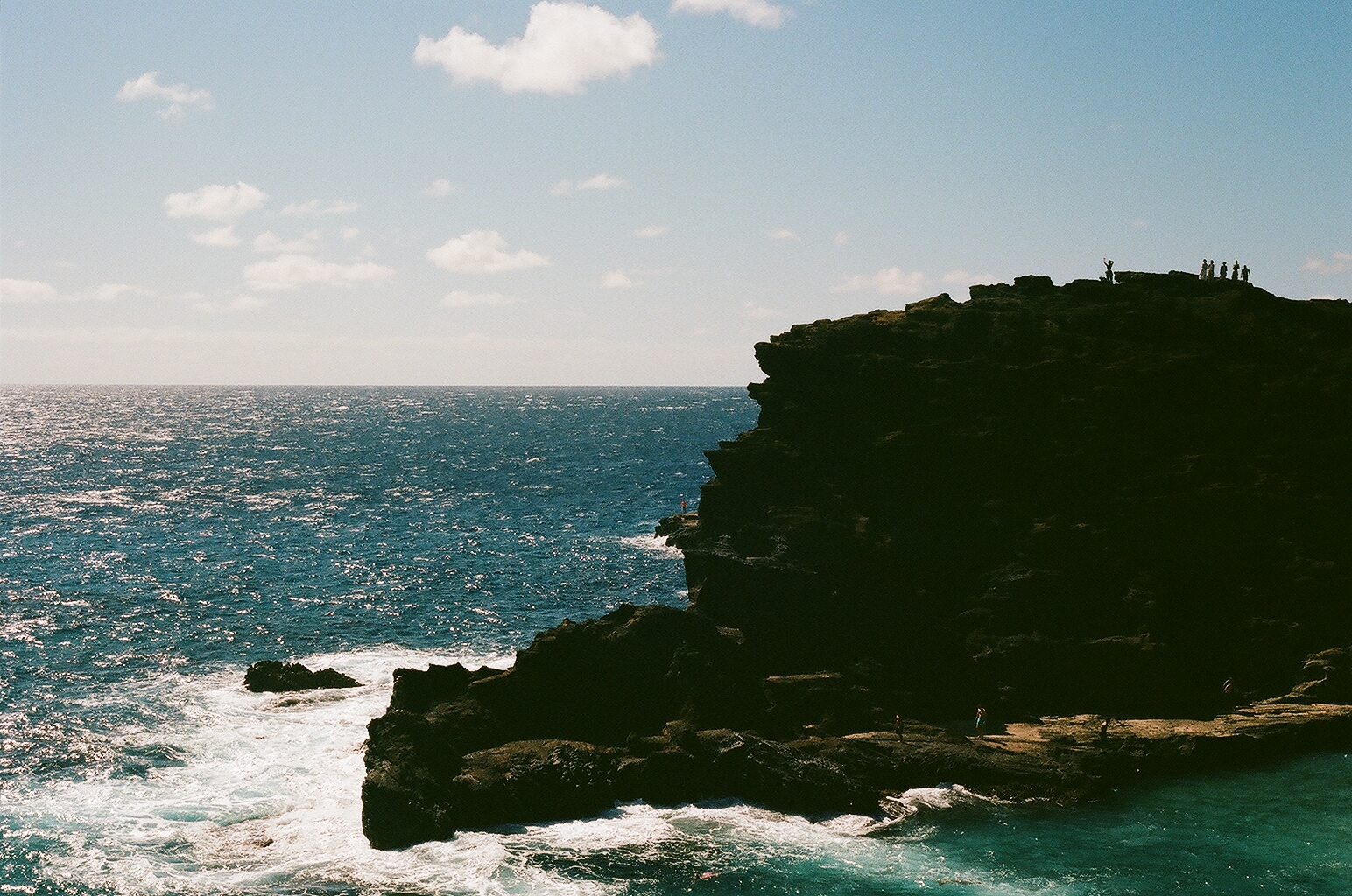 Tiny figures stand atop a shadowed lava headland above the glittering open ocean