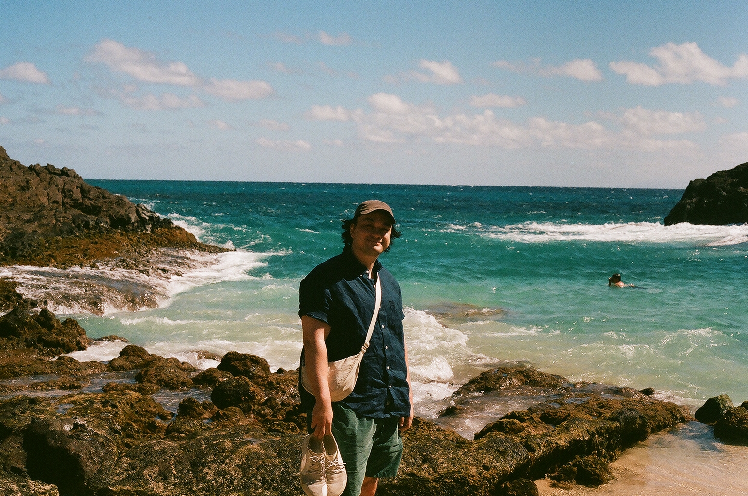 A smiling man in a cap holds his sneakers on lava rocks beside turquoise surf