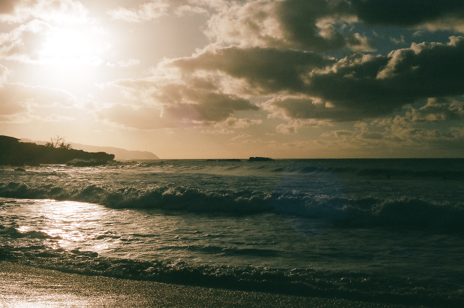 Low sun breaking through heavy clouds over dark surf and a silhouetted headland