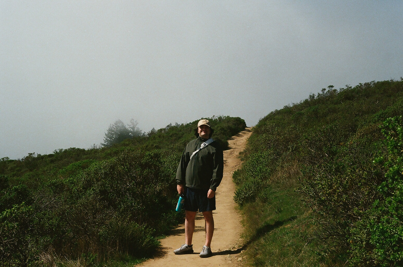 A man in a cap and shorts pausing on a dirt trail up a foggy hillside