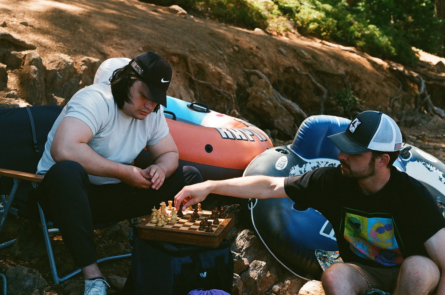 Two friends in ball caps playing chess on a rocky shore beside inflatable rafts