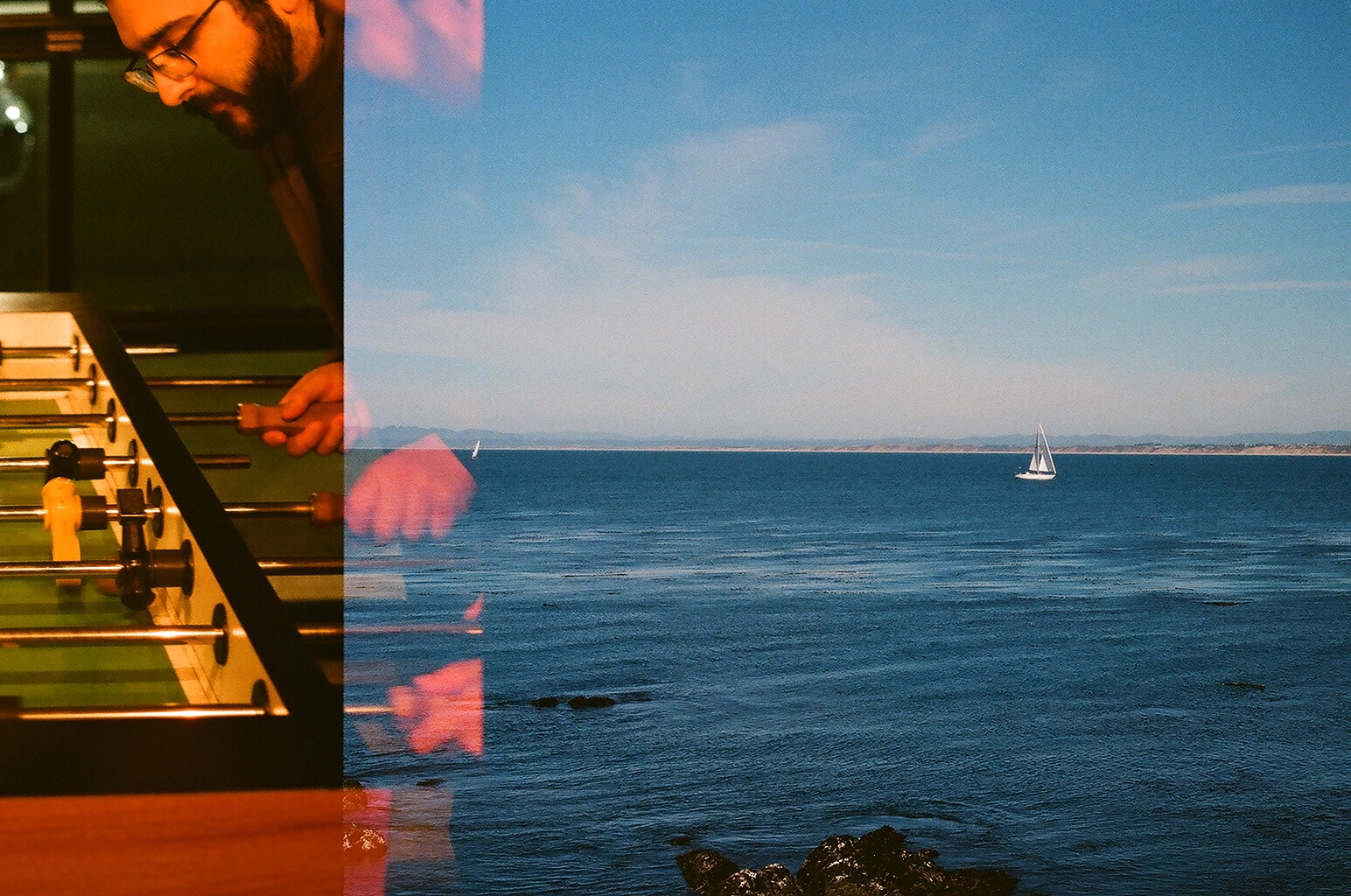 Double exposure of a bearded man playing foosball and a sailboat on calm open water