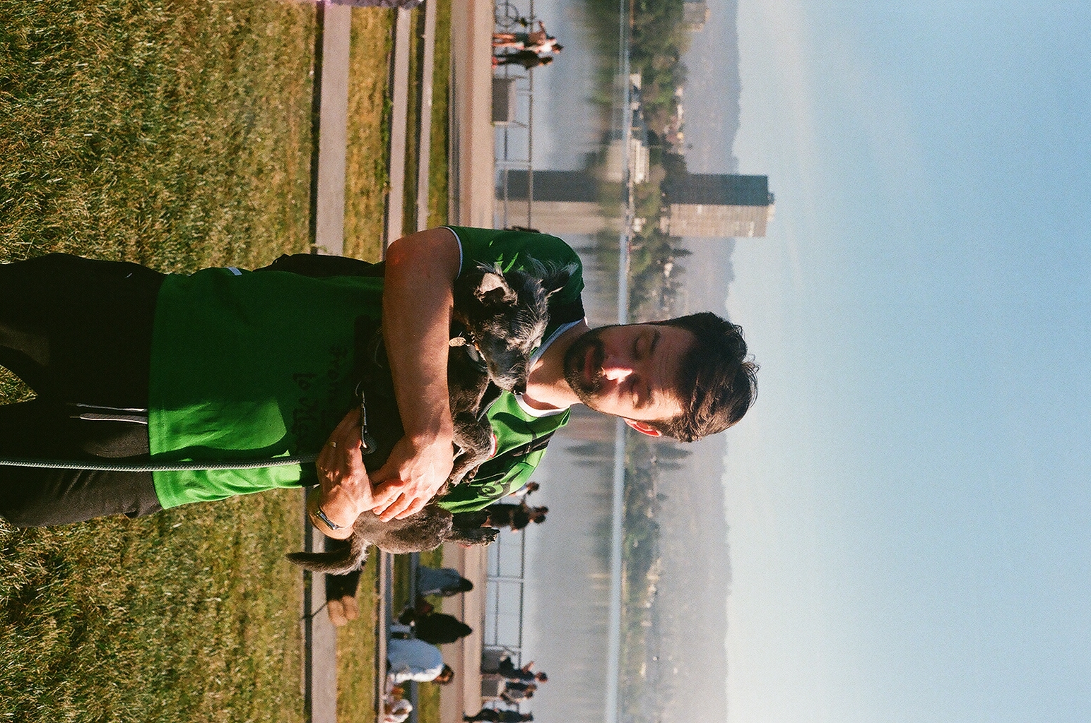 A man in a green shirt cradles a small scruffy black dog beside Lake Merritt