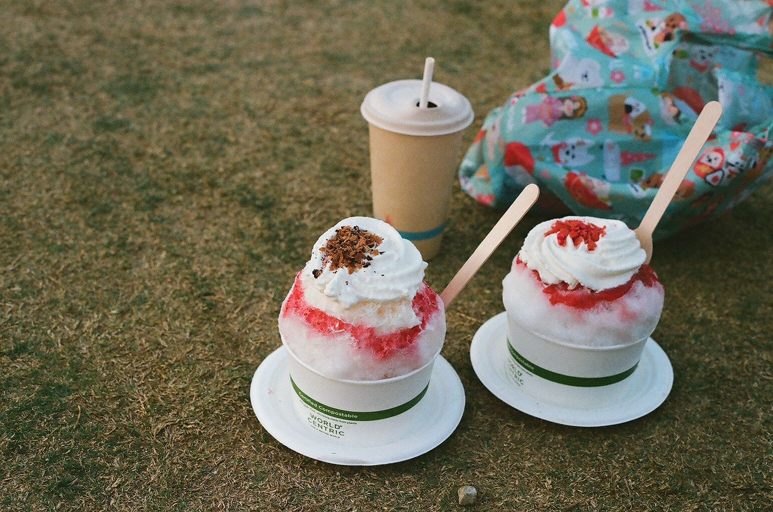 Two cups of shaved ice topped with whipped cream on the grass beside an iced drink